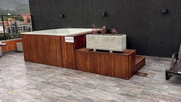 Una terraza en la azotea con un banco de madera, una jardinera de concreto con plantas y una parrilla de metal.