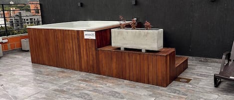 Una terraza en la azotea con un banco de madera, una jardinera de concreto con plantas y una parrilla de metal.