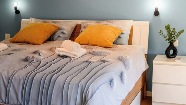 Een bed met een blauw-witte spreidek, twee gele kussens en een witte nachtkastje met een plant.