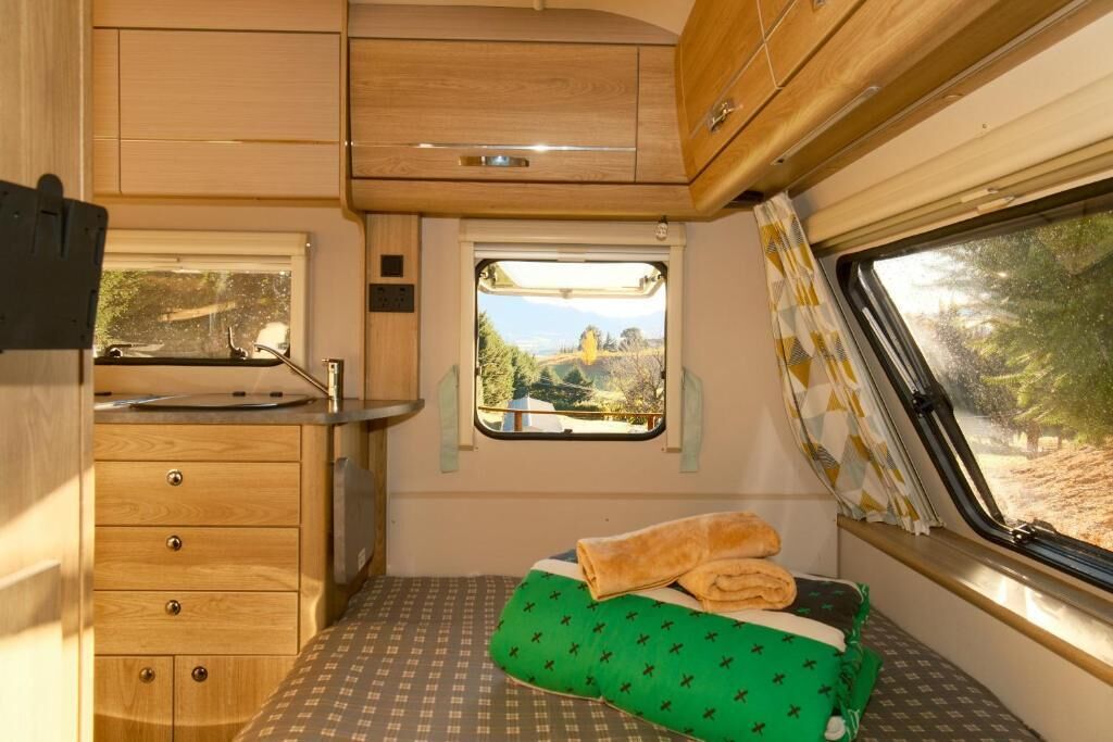 Interior de una caravana compacta con kitchenette, una pequeña ventana con vistas, cama con colcha estampada verde y armarios de madera.