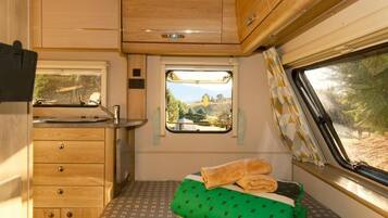 Interior de una caravana compacta con kitchenette, una pequeña ventana con vistas, cama con colcha estampada verde y armarios de madera.