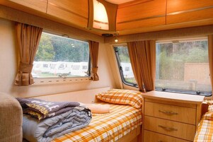Interior de una caravana compacta con cama, cómoda pequeña y dos ventanas.