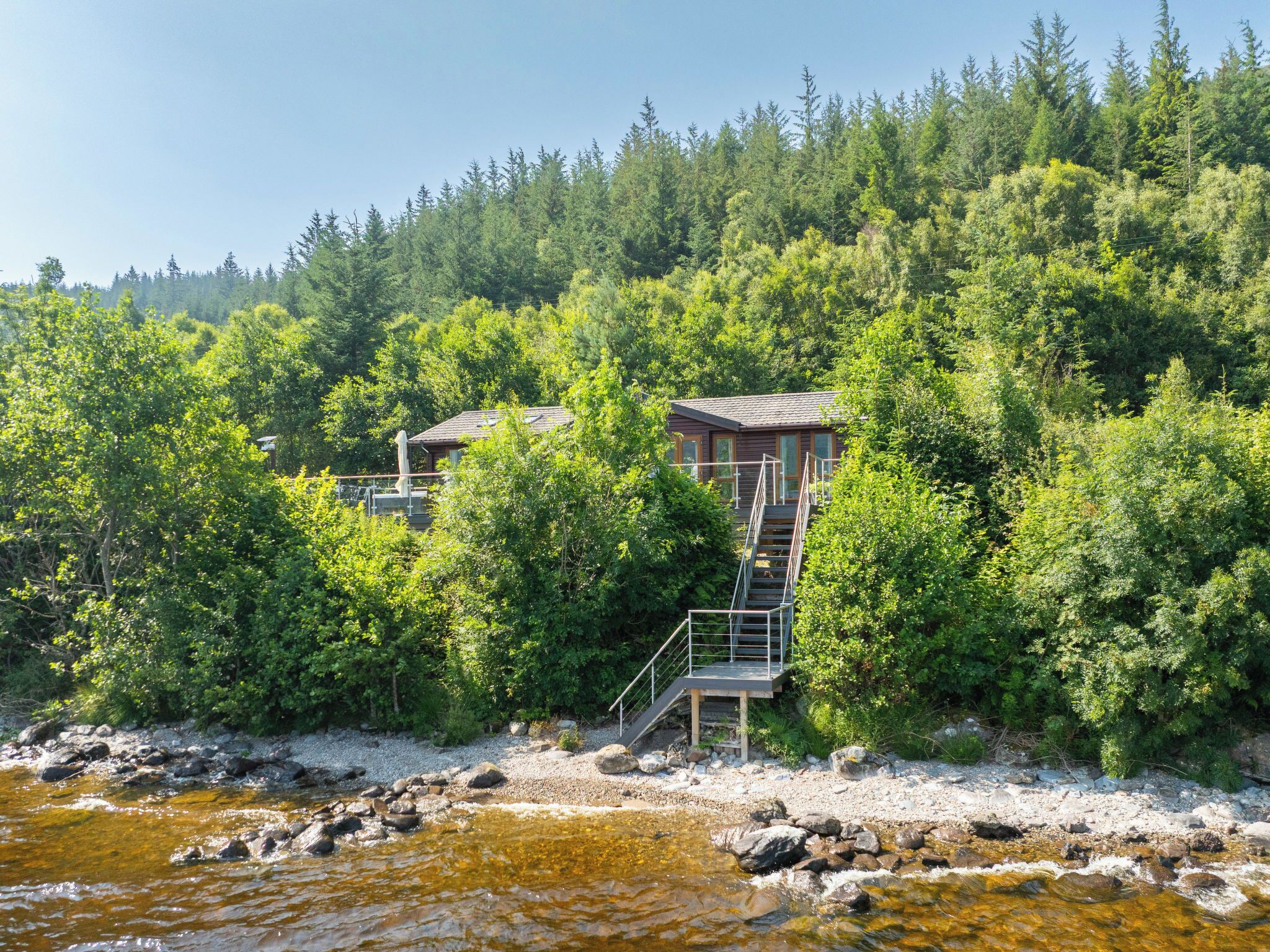 Eine Holzhütte mit einer Treppe, die zu einer Terrasse führt, umgeben von dichtem Wald und einem Fluss.