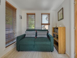 Un canapé vert agrémenté de coussins à motifs, des stores en bois aux fenêtres, une bibliothèque et un tableau encadré au mur.