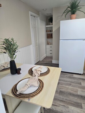 Eine Küche mit einem weißen Kühlschrank, einem Holztisch mit Platzsets, einer Topfpflanze und einer weißen Tür.