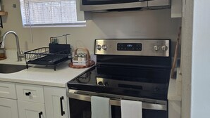 Ett kök med vita skåp, en mikrovågsugn i rostfritt stål, en spis och ett fönster med persienner.