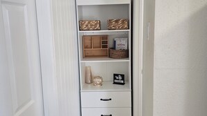 En smal hall med en vit dörr på vänster sida, en vit hylla med korgar och en skylt, och en växt ovanpå.