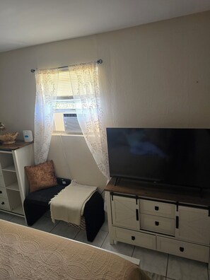 A small room with a bed, a TV on a stand, a window with a curtain, and a white cabinet with drawers. - Cozy 1-bedroom studio in charming Sarasota. (Sarasota)