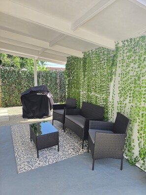 Eine Terrasse im Freien mit Korbmöbeln, einem Grill und einem gemusterten Teppich.
