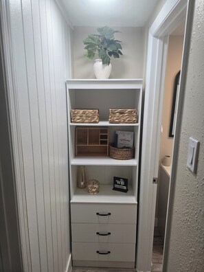 A narrow hallway with a white shelf unit containing baskets, a welcome sign, a WiFi sign, and a potted plant. - Cozy 1-bedroom studio in charming Sarasota. (Sarasota)