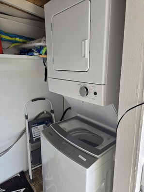 Un espace buanderie compact comprenant un lave-linge et un sèche-linge à chargement frontal, une petite étagère avec des vêtements pliés et un chariot muni d’un câble d’alimentation.