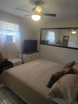Une chambre à coucher comprenant un lit, un téléviseur, un ventilateur de plafond, un miroir et une fenêtre avec des rideaux.