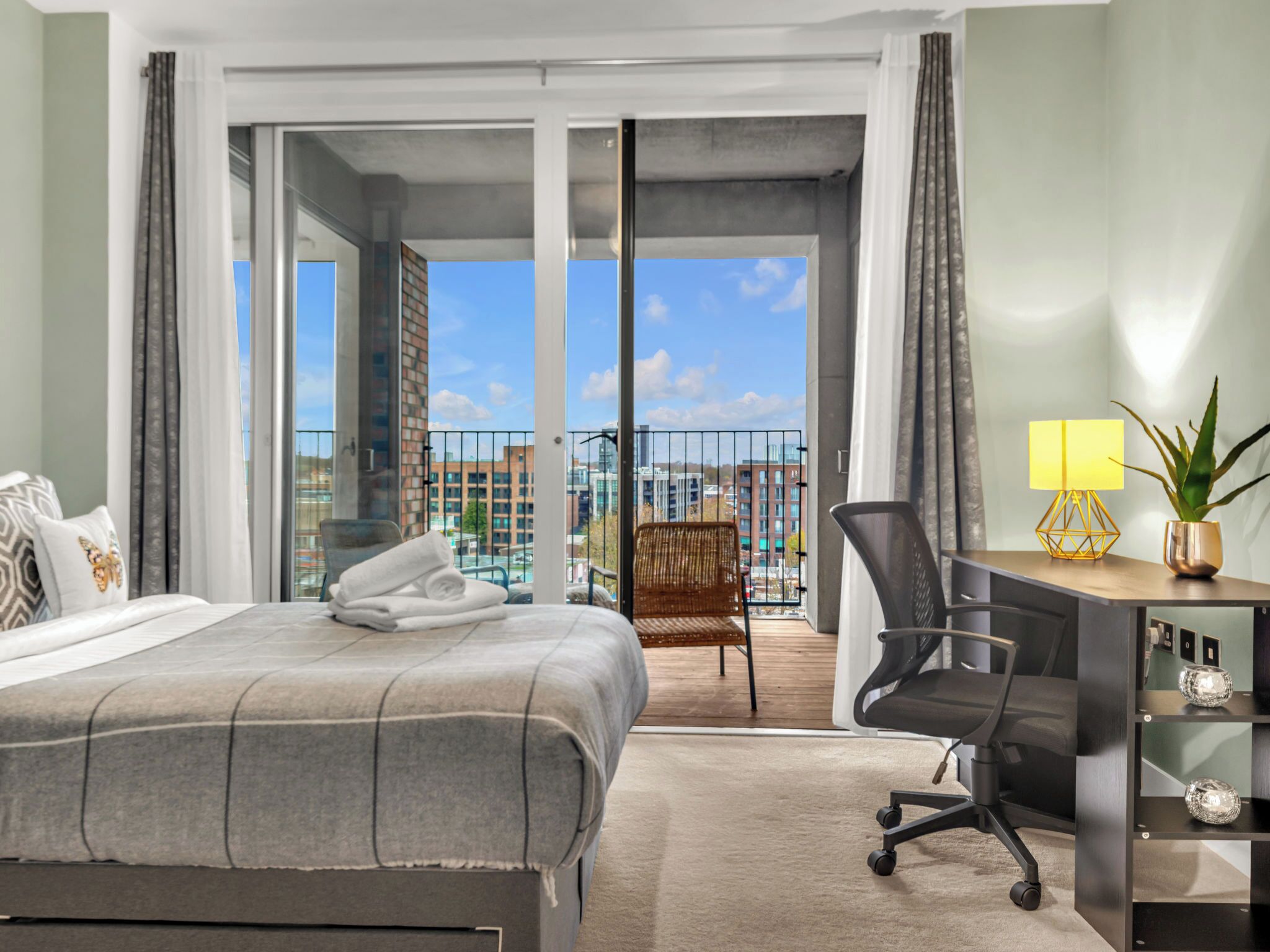 A hotel room with a bed, a desk, a chair, a balcony with a view of the city, and a plant.
