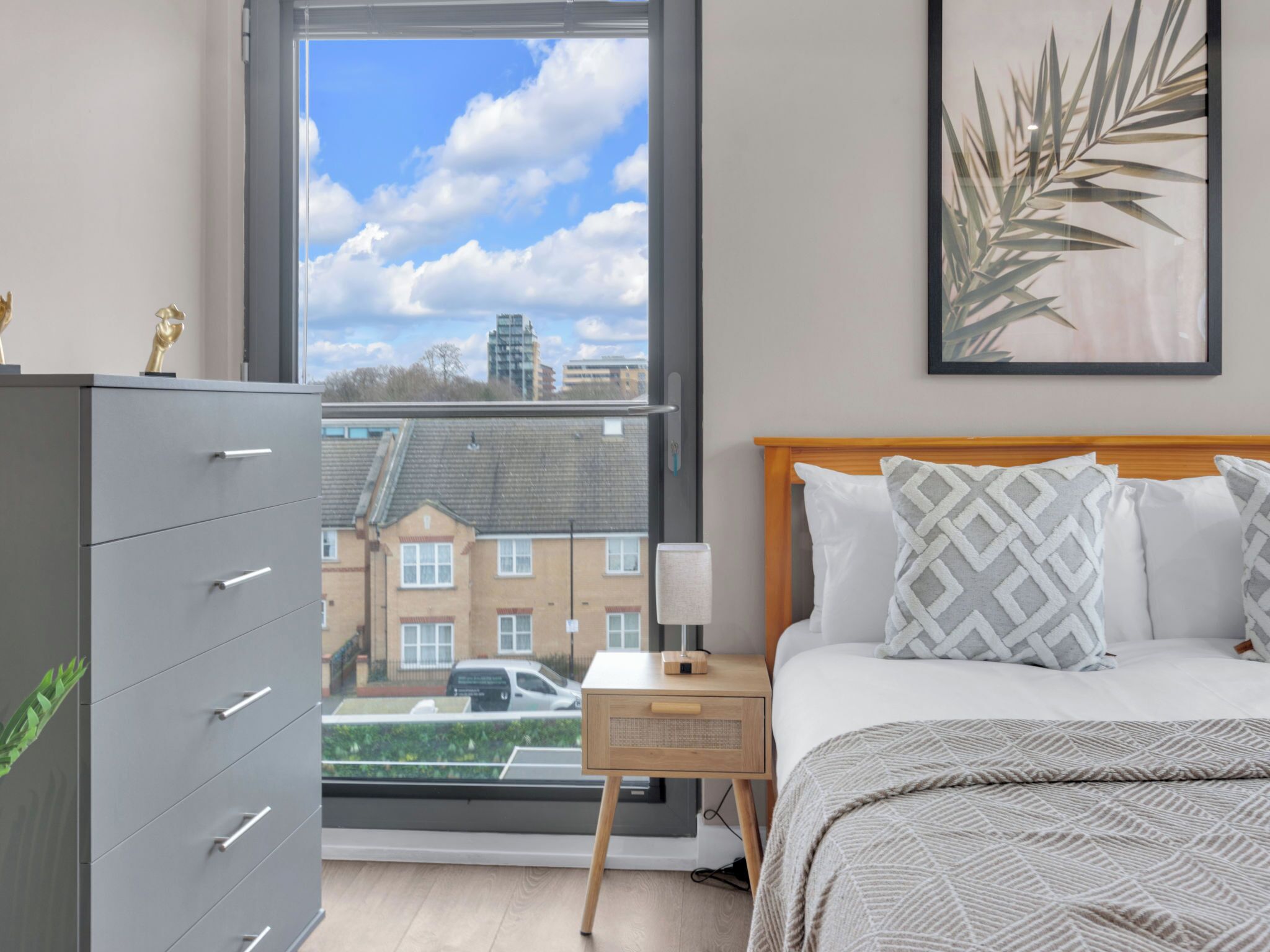 Un dormitorio con cama, mesita de noche, cómoda, ventana con vistas a edificios y una planta enmarcada en la pared.