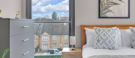 A bedroom with a bed, a nightstand, a dresser, a window with a view of buildings, and a framed plant on the wall.