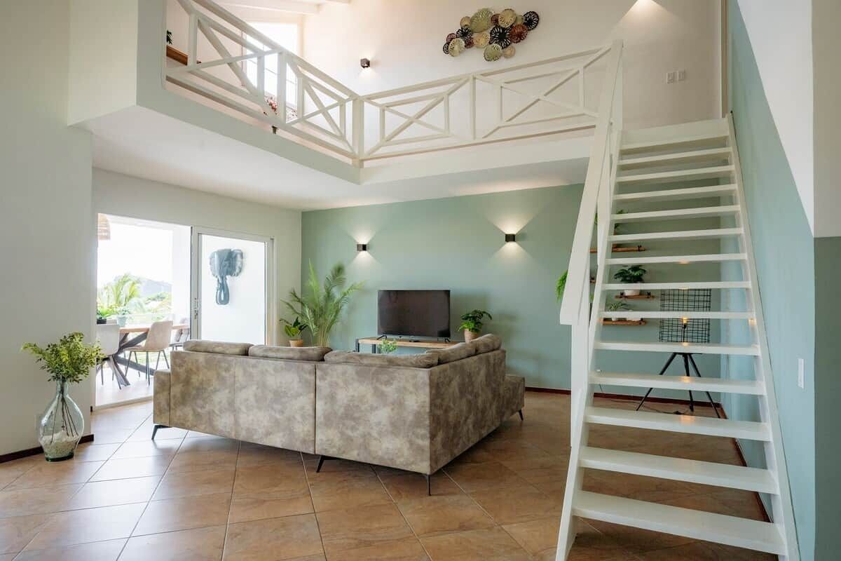 A modern living room with a sofa, TV, and plants. There's a staircase with a railing and a balcony with a railing.