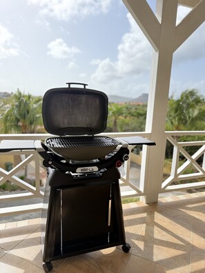 Un barbecue Weber portable sur un balcon, avec vue sur des palmiers et des collines lointaines.