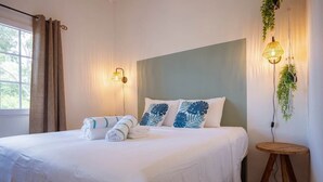 Una cama bien hecha con sábanas blancas y almohadas estampadas con hojas azules, una lámpara de noche y un pequeño taburete de madera.
