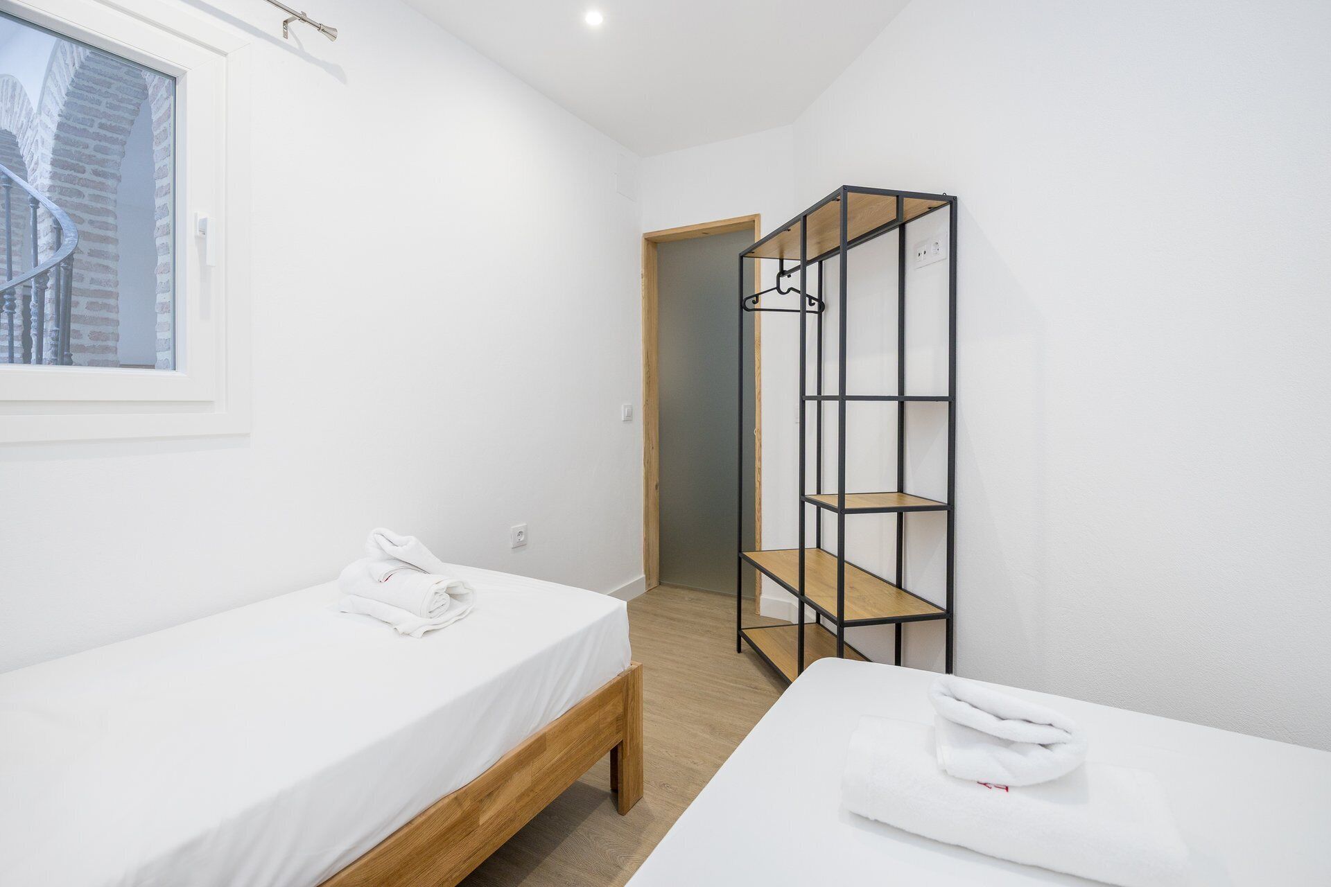Una habitación con dos camas, un armario de madera, un ventanal con vista a un edificio de ladrillo y una puerta que da a otra habitación.
