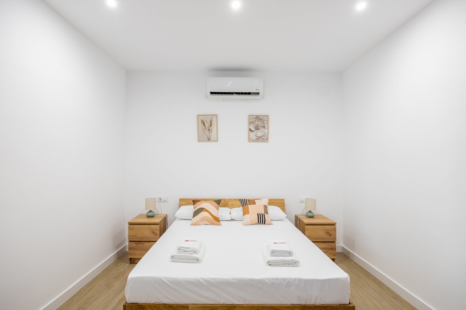A hotel room with a bed, two bedside tables, an air conditioning unit, and two wall-mounted pictures.