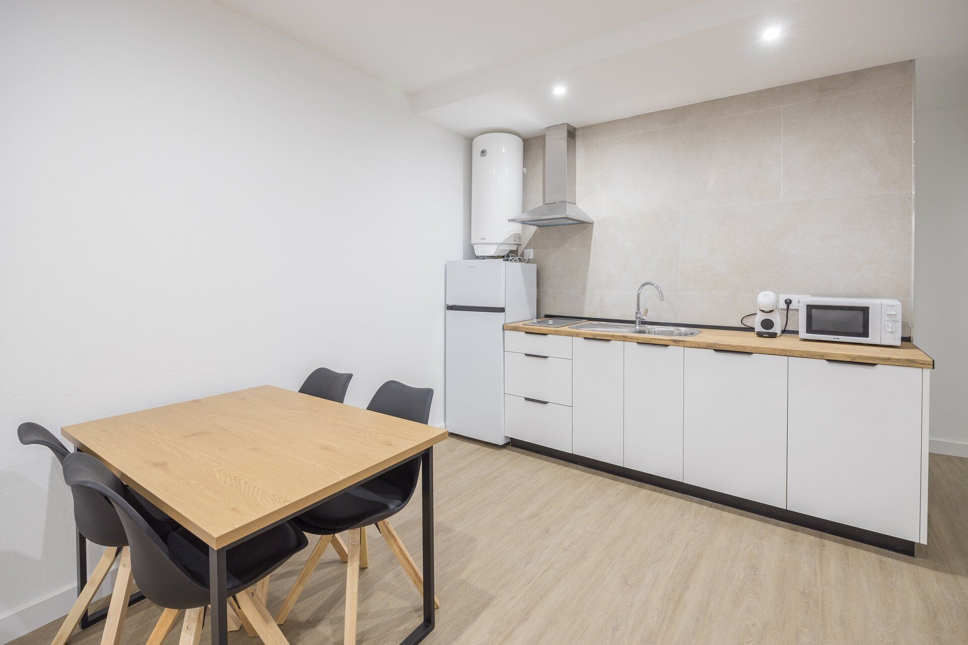 Cocina compacta con una mesa de madera, dos sillas, microondas, hervidor y horno empotrado.