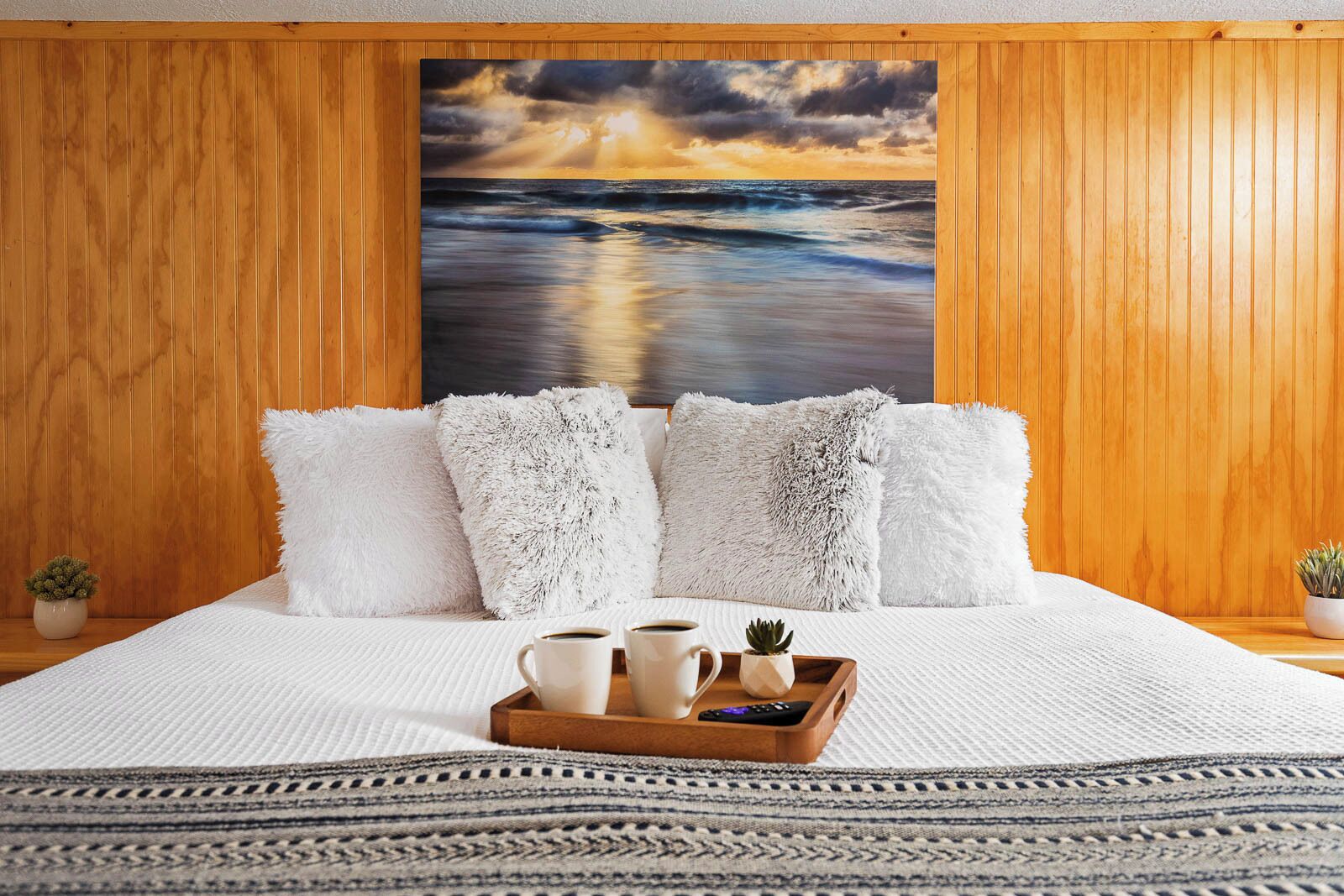 A hotel room with a large painting of a sunset over the ocean, fluffy white pillows, a tray with coffee cups, and a small potted plant.