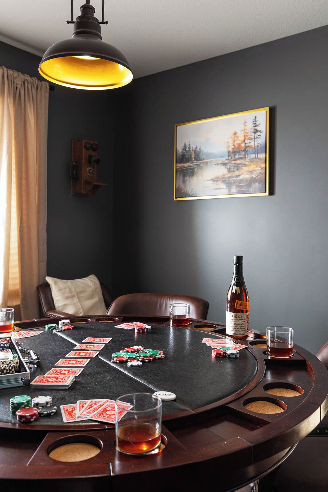 A room with a poker table, chairs, a bottle, glasses, and a painting on the wall.