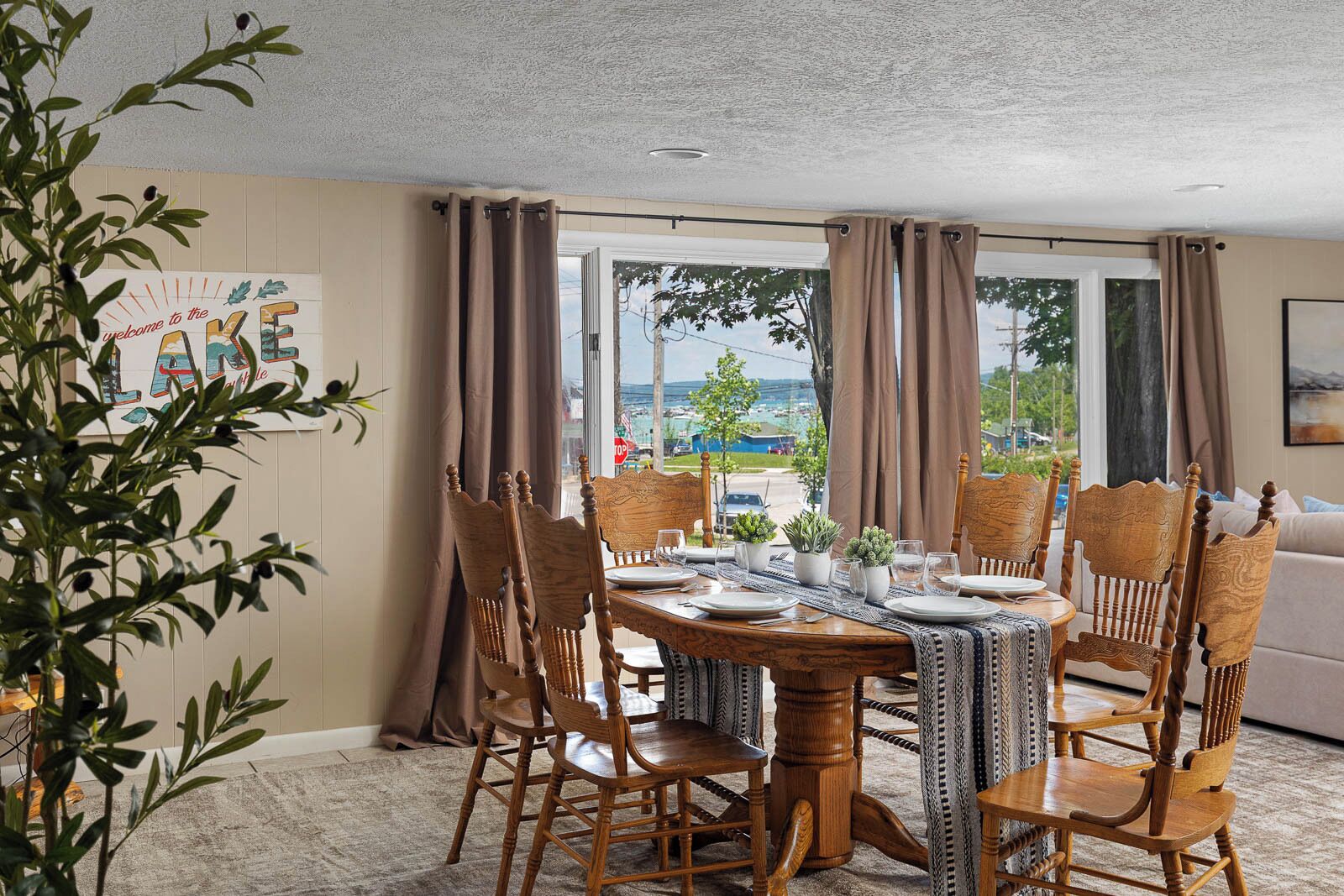 A dining area with a table set for four, surrounded by wooden chairs, with a view of a street and greenery outside.