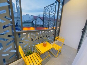 Un balcón con sillas metálicas amarillas y una mesa, una planta en maceta, y vistas de edificios al fondo.