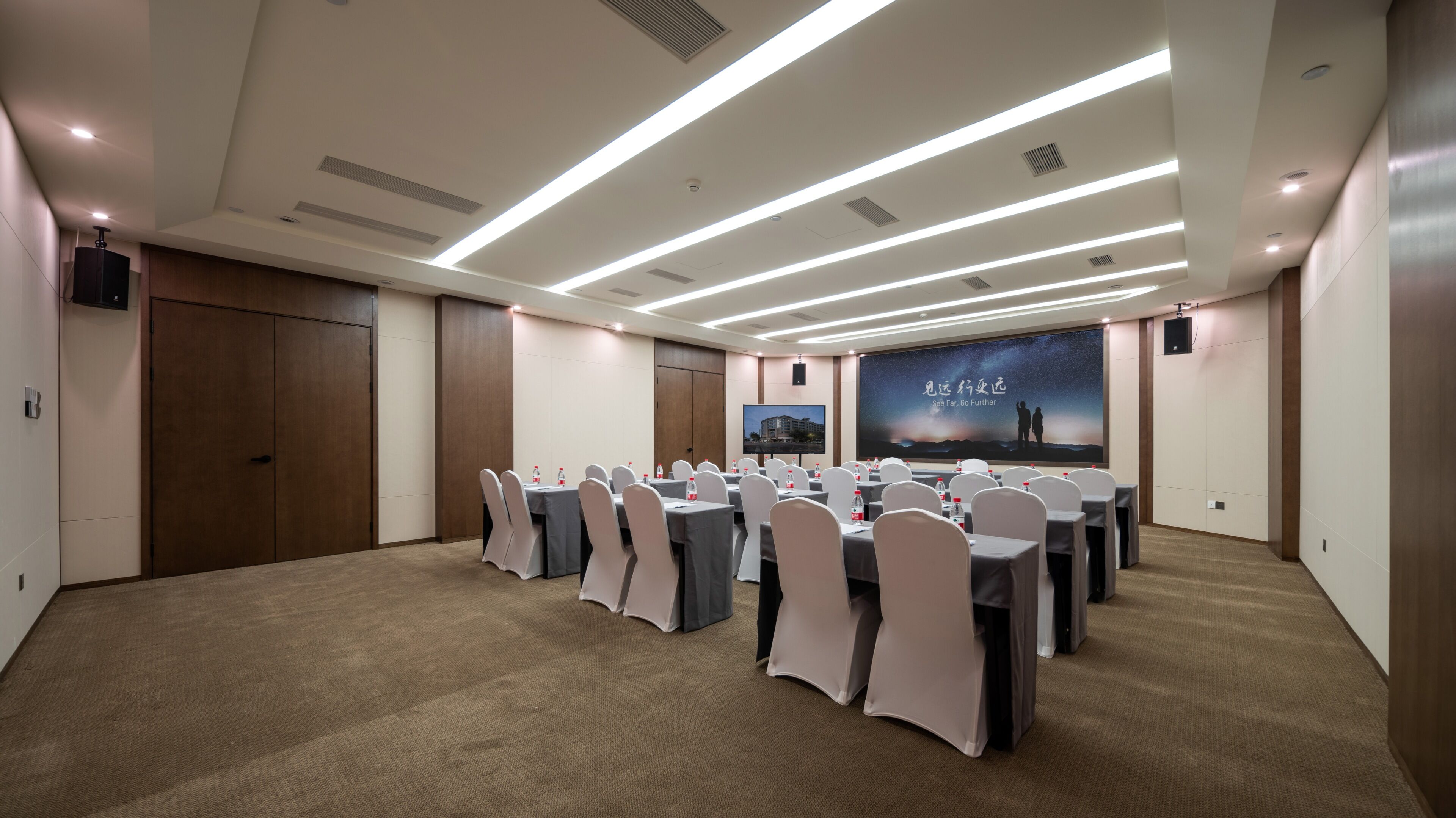 Un salón de conferencias con mesas dispuestas para una reunión, una pantalla grande mostrando una presentación y una puerta a la izquierda.