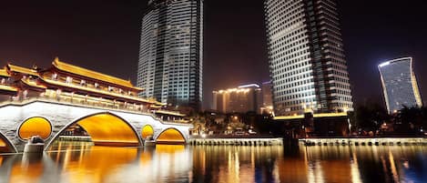 A night view of a cityscape with modern high-rise buildings and a traditional bridge with illuminated arches.