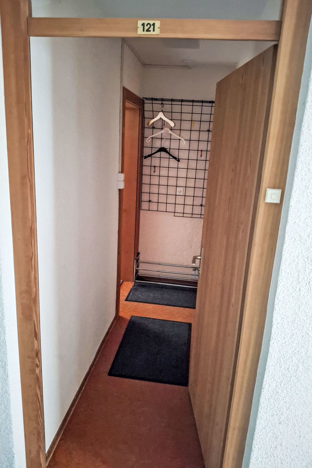 Ein schmaler Korridor mit einem hölzernen Türrahmen, einem Schild mit der Nummer 121, einem Metallgestell mit Kleiderbügeln und einer schwarzen Matte auf dem Boden.