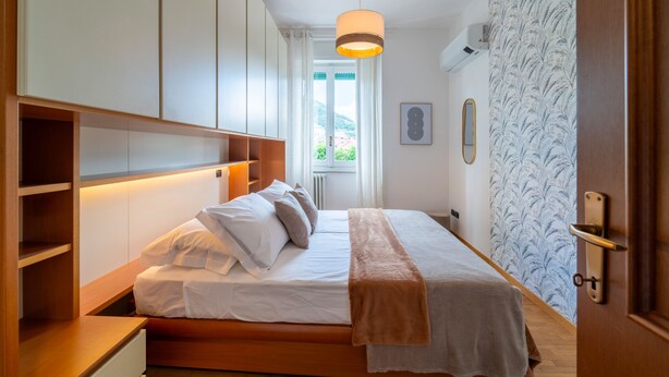 A bedroom with a bed, wooden headboard, bedside tables, a window with curtains, and a wall-mounted air conditioning unit. - Manzoni 2 - Beyond the Duomo (Como)