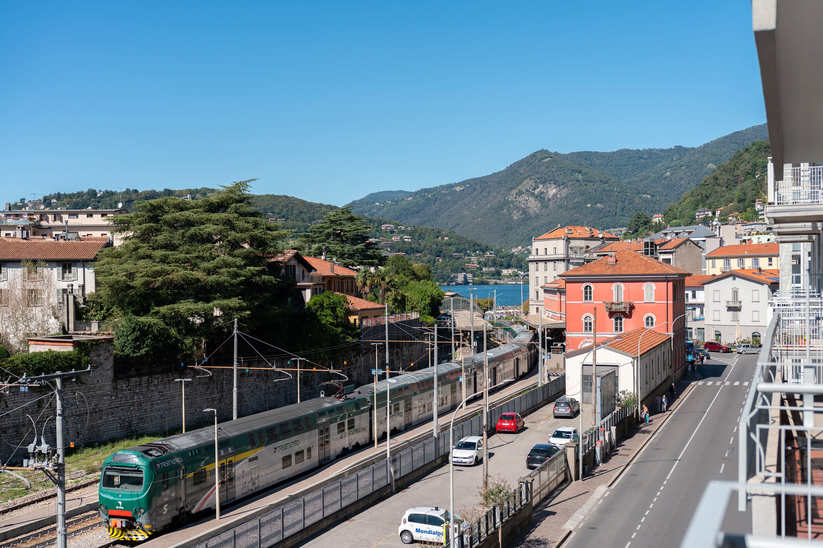 Una estación de tren con un tren verde, una carretera con coches y edificios al fondo.