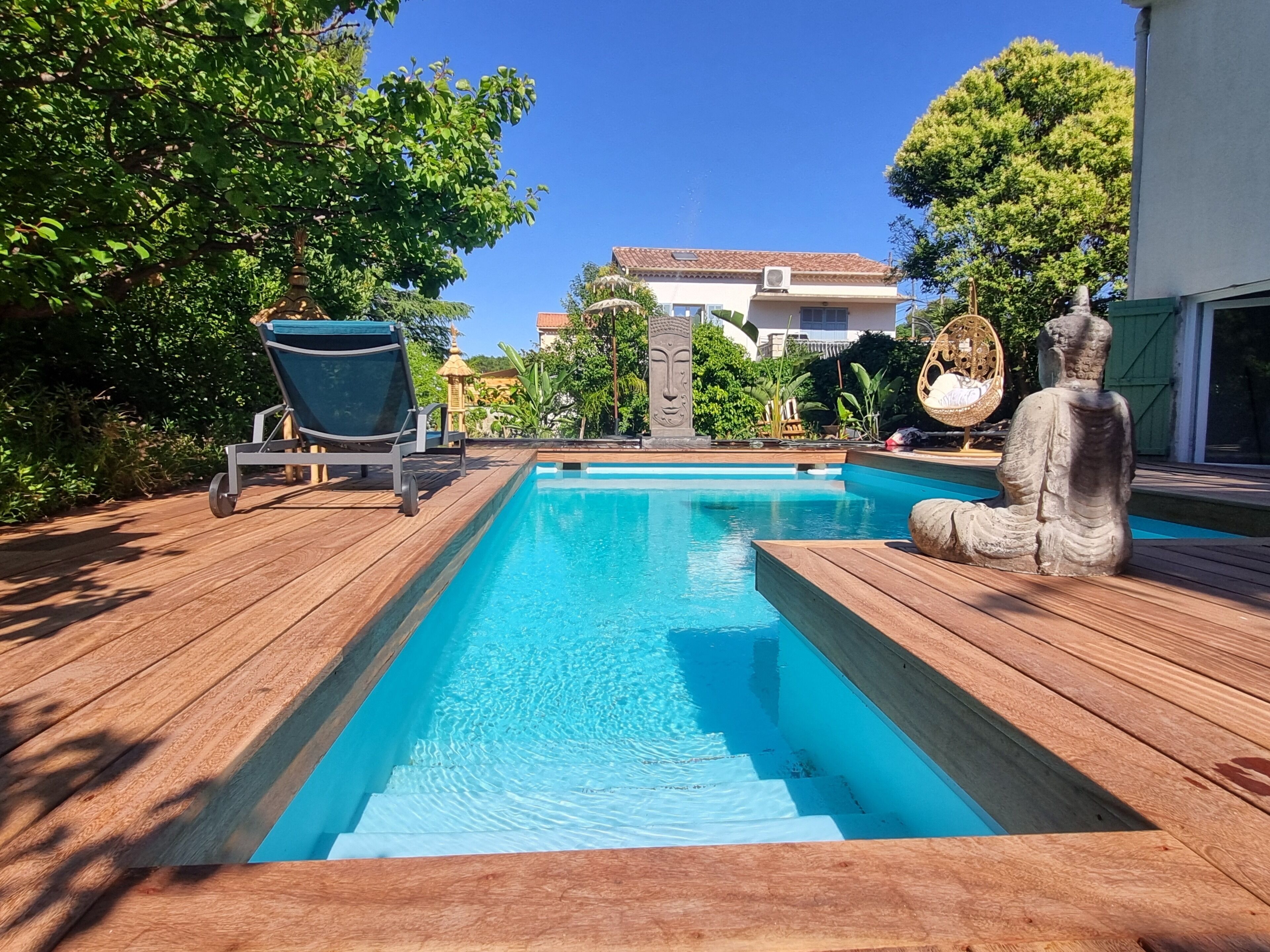 A rectangular swimming pool with a wooden deck, a lounge chair, a statue, and a building in the background.