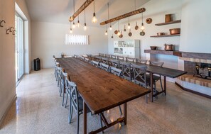 Une longue table en bois avec des chaises, un coin cuisine avec des étagères et des luminaires suspendus.