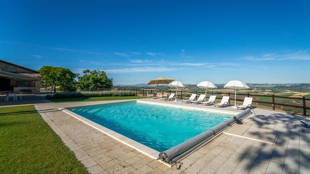 Una piscina con sillas de descanso y sombrillas, rodeada por una terraza de madera y vistas al campo.