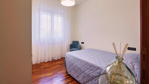 Un dormitorio con cama, una silla, ventana con cortinas y un florero con ramas de juncos.