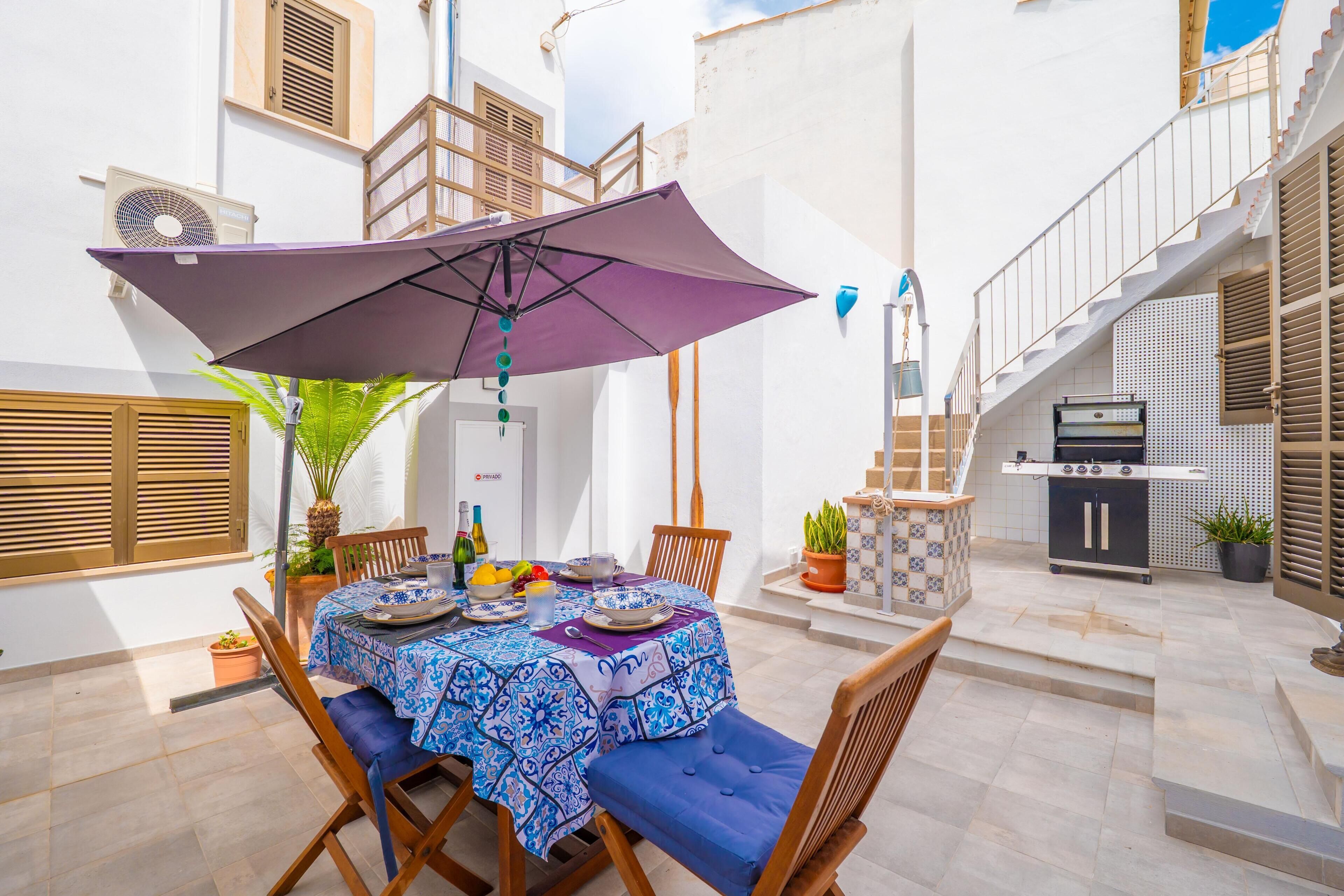 An outdoor dining area with a table set for two, surrounded by wooden chairs, under a purple umbrella.