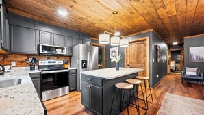 A modern kitchen with dark cabinets, stainless steel appliances, a central island with stools, and a wooden ceiling. - 3 King Suites, Game Room, Hot Tub - 2 Mi to Island (Pigeon Forge)