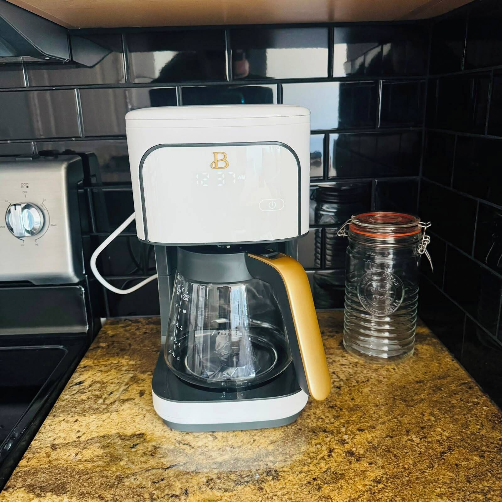 Une machine à café dotée d’un écran numérique, d’une verseuse en verre et d’un récipient en acier inoxydable, posée sur un plan de travail de cuisine.