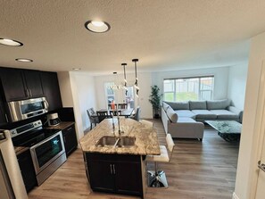 A modern kitchen with a central island, dark cabinetry, stainless steel appliances, and a dining area with a table and chairs. - Modern Windermere Home | Sleeps 6 | Near Shopping & Airport (Edmonton)
