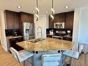 A modern kitchen with dark wood cabinets, stainless steel appliances, a granite countertop, and bar stools. - Modern Windermere Home | Sleeps 6 | Near Shopping & Airport (Edmonton)
