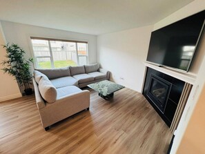 Een woonkamer met een hoekbank, een flatscreen televisie boven een open haard, en een potplant.