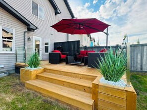 A wooden deck with outdoor furniture, a red umbrella, and a planter with green plants. - Modern Windermere Home | Sleeps 6 | Near Shopping & Airport (Edmonton)