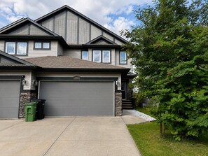 A two-story house with a grey garage door, stone accents, and a well-maintained lawn. - Modern Windermere Home | Sleeps 6 | Near Shopping & Airport (Edmonton)