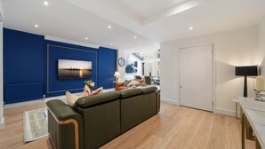 A modern living room with a sectional sofa, a flat-screen TV, a clock, and a dining area with a table and chairs. - Superior Bedroom Apartment In Mayfair (London)