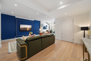 A modern living room with a sectional sofa, a flat-screen TV, a clock, and a dining area with a table and chairs. - Superior Bedroom Apartment In Mayfair (London)