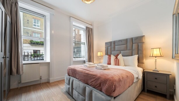 A hotel room with a large bed, two bedside lamps, a nightstand, and a view of the city through the windows. - Superior Bedroom Apartment In Mayfair (London)
