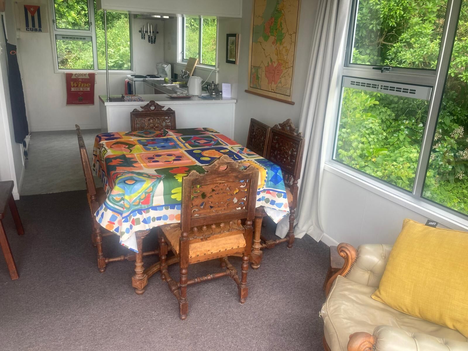 A dining area with a table covered by a colorful quilt, wooden chairs, a window with a view of greenery, and a map on the wall.